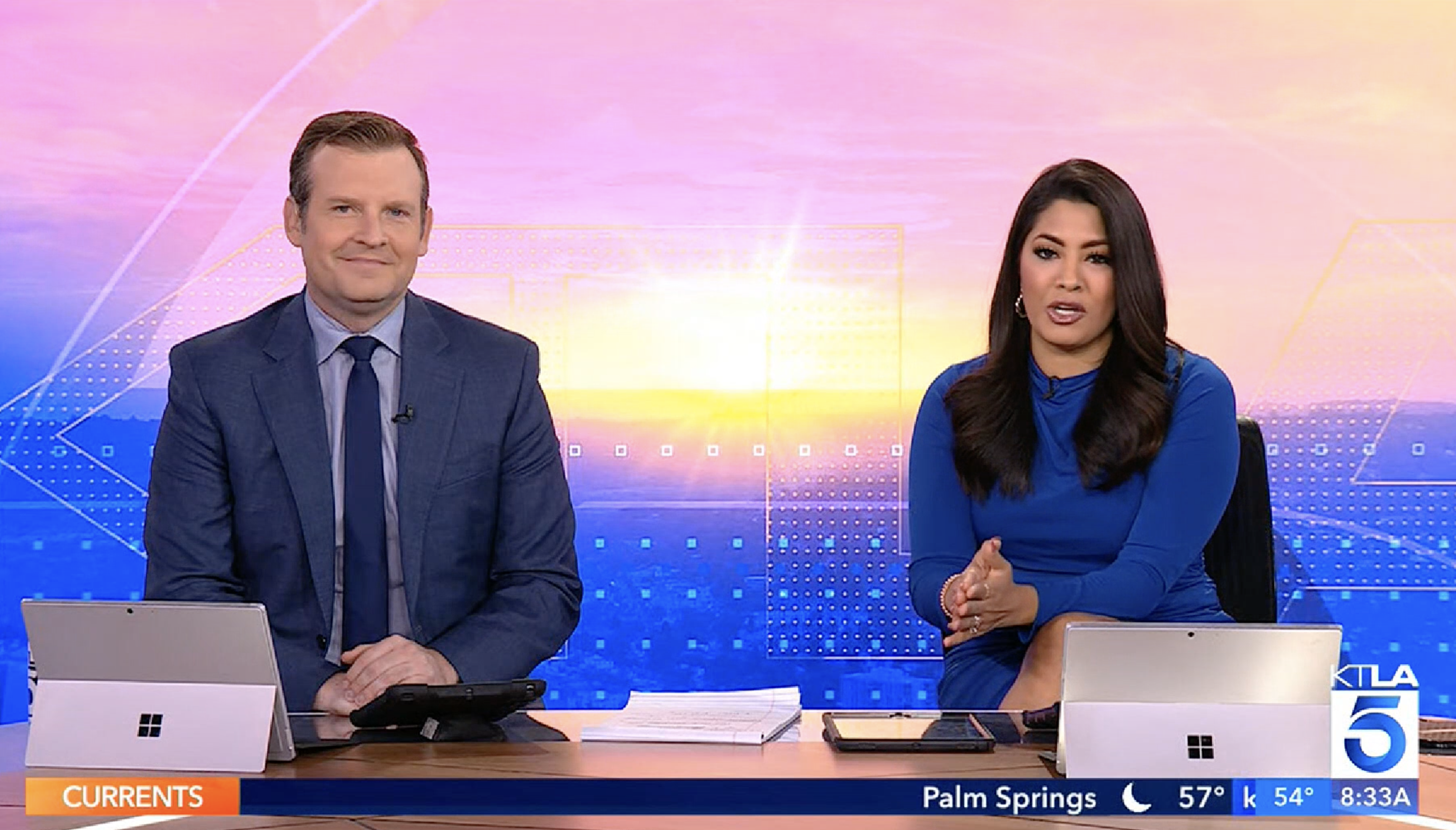 Two news anchors presenting on a television broadcast with a cityscape in the background.